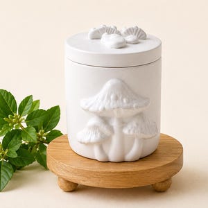 May include: A white cylindrical ceramic jar with a lid featuring three sculpted mushroom figures on the side and three small sculpted mushroom caps on the top. The jar is placed on a round, light-coloured wooden stand with three short, rounded feet.