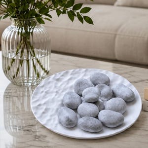May include: A white, round, textured decorative bowl filled with a collection of smooth, grey, oval-shaped river stones. The bowl features an uneven, rippled surface texture, and the stones vary slightly in size and shade of grey.