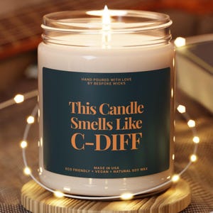 May include: A lit candle in a clear glass jar with a teal label. The label reads "This Candle Smells Like C-DIFF" in orange text. The candle is made with natural soy wax and is eco-friendly.