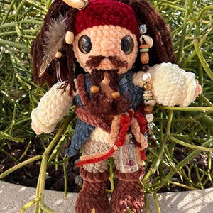 Könnte beinhalten: Eine gehäkelte Puppe mit dunkelbraunem Garnhaar, einem roten Stirnband und einem Schnurrbart. Die Puppe trägt eine blaue Weste, cremefarbene Ärmel und braune Stiefel. Zu den Accessoires gehören eine kleine Glasflasche, Perlen und ein weißes Horn.