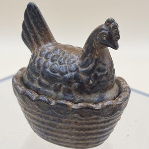 May include: A dark brown ceramic figurine of a hen sitting in a basket. The hen has detailed moulded feathers and a prominent comb on its head. The basket base features a scalloped rim and a textured, woven-like pattern across its exterior surface.