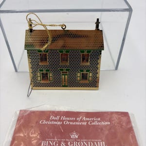 Può includere: Un ornamento a forma di casa in metallo dorato con motivo a mattoni, accenti verdi e un cordino dorato per appenderlo. Sotto l'ornamento c'è un inserto di carta rossa con l'etichetta Doll Houses of America Christmas Ornament Collection di Bing & Grøndahl.