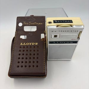 以下が含まれることがあります： クリーム色のトップとシルバーの金属製スピーカーグリルを備えたヴィンテージのLloyd's製6トランジスタラジオ。「SIX TRANSISTOR」と「LLOYD'S」の文字が記されています。横には「LLOYD'S」と「Ira Gastman」とプリントされた茶色のレザー製キャリングケースが置かれています。