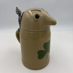 May include: A tan ceramic beer stein featuring a lid shaped like a bird head with a prominent beak and dark eyes. The cylindrical body is decorated with a green shamrock graphic and includes a dark metal handle with an ornate thumb lever.