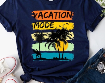 以下が含まれることがあります： オレンジと黄色で「VACATION MODE」の文字が入ったネイビーブルーのTシャツ。ビーチシーンのグラフィックが描かれています。ヤシの木、サーファー、夕焼けが含まれています。白いスニーカーと青いジーンズがシャツの隣にあります。