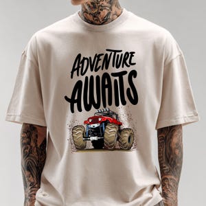 Peut inclure: T-shirt beige avec l'inscription noire "ADVENTURE AWAITS" au-dessus d'une illustration de monster truck rouge. Le camion a de gros pneus et projette de la terre. Le t-shirt est de style d&eacute;contract&eacute;.