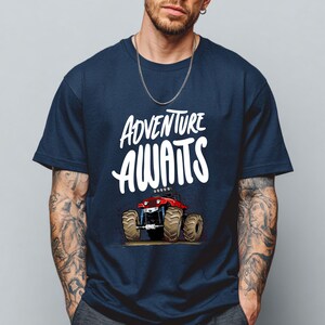 Peut inclure: T-shirt bleu marine avec l'inscription blanche "ADVENTURE AWAITS" au-dessus d'un dessin de monster truck rouge. Le camion a de gros pneus bruns et se d&eacute;tache sur un fond brun clair.