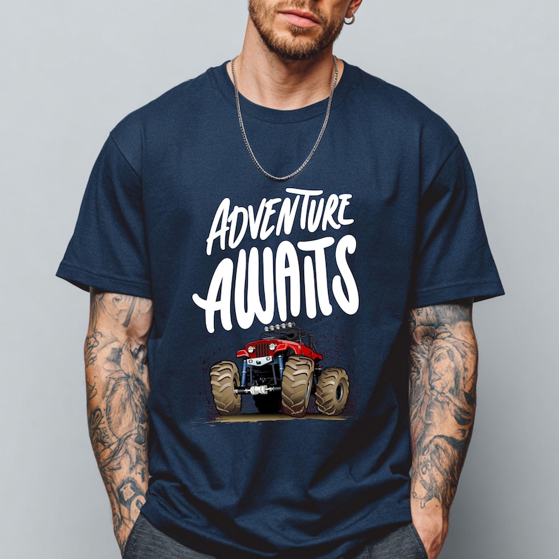 Peut inclure: T-shirt bleu marine avec l'inscription blanche "ADVENTURE AWAITS" au-dessus d'un dessin de monster truck rouge. Le camion a de gros pneus bruns et se d&eacute;tache sur un fond brun clair.