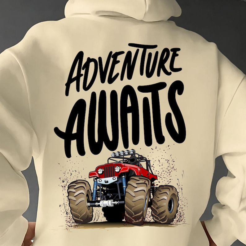 Peut inclure: Sweat &agrave; capuche cr&egrave;me avec un motif de monster truck rouge et les mots "ADVENTURE AWAITS" en noir. Le camion a de gros pneus et une suspension bleue. Le design comprend des &eacute;claboussures de terre.