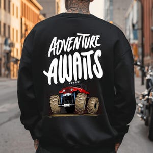 Peut inclure: Sweat-shirt noir avec l'inscription blanche "ADVENTURE AWAITS". En dessous, un dessin de monster truck rouge avec de gros pneus. Le sweat-shirt est de style col rond.