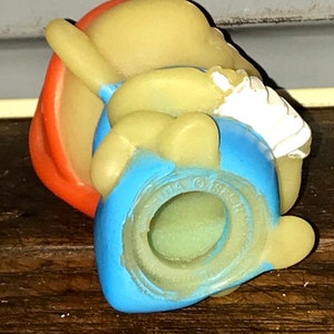 Puede incluir: Una figurita de goma amarilla de un personaje de dibujos animados que lleva un sombrero rojo, pantalones azules y una camisa blanca. La figurita est&aacute; sentada con las piernas cruzadas y los brazos cruzados. La figurita tiene un agujero en la parte inferior, lo que sugiere que puede ser un juguete o una decoraci&oacute;n.