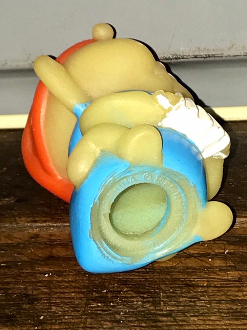 Puede incluir: Una figurita de goma amarilla de un personaje de dibujos animados que lleva un sombrero rojo, pantalones azules y una camisa blanca. La figurita est&aacute; sentada con las piernas cruzadas y los brazos cruzados. La figurita tiene un agujero en la parte inferior, lo que sugiere que puede ser un juguete o una decoraci&oacute;n.