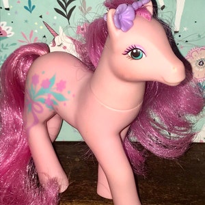 Puede incluir: Pony My Little Pony rosa con una corona de flores moradas y un dise&ntilde;o floral en un lado. El pony tiene el pelo rosa y los ojos azules.