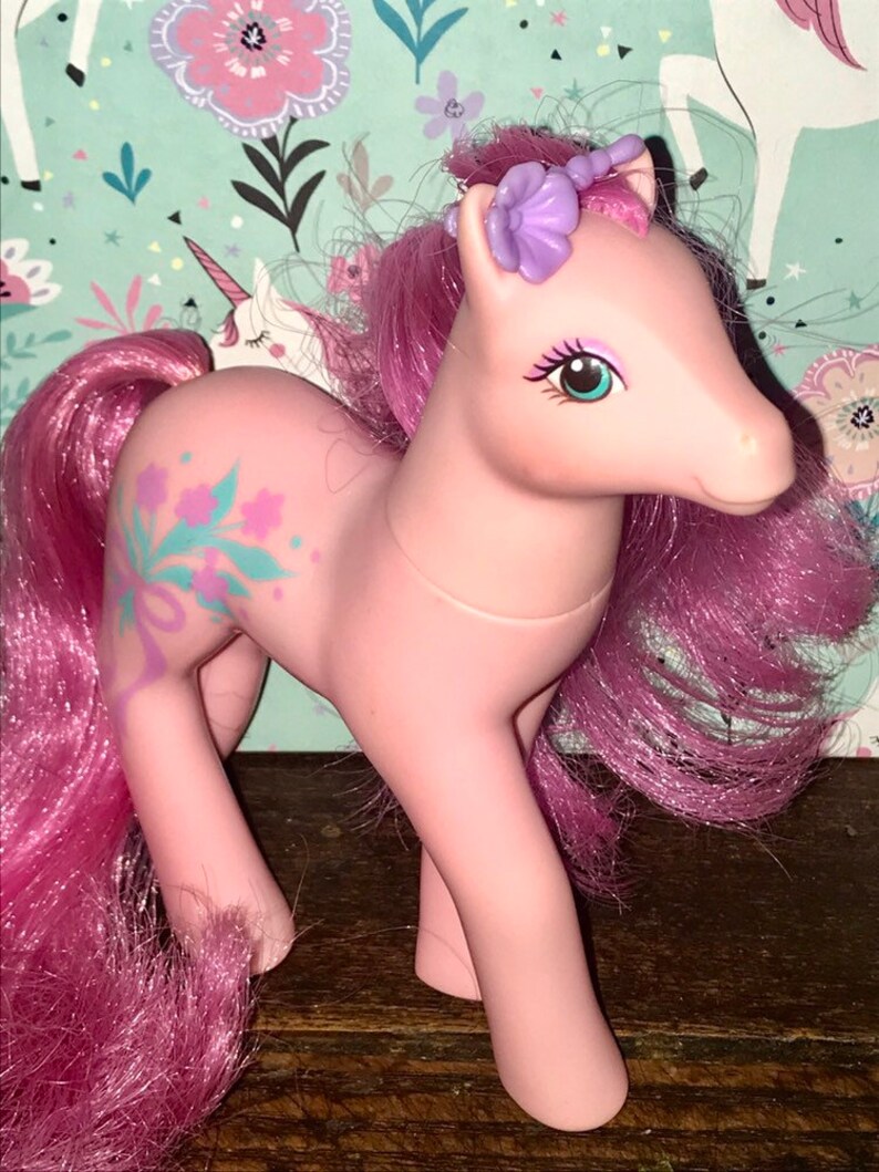 Puede incluir: Pony My Little Pony rosa con una corona de flores moradas y un dise&ntilde;o floral en un lado. El pony tiene el pelo rosa y los ojos azules.