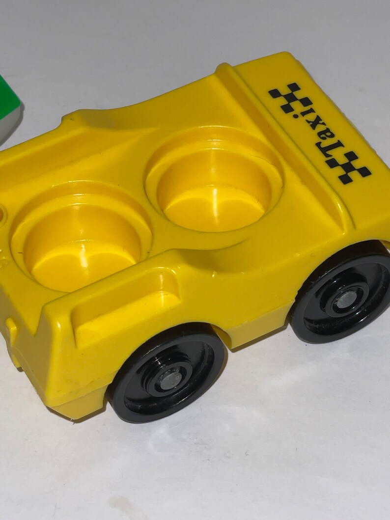 May include: Yellow plastic toy taxi with black wheels and two empty cup holders. The taxi has a black and white checkered design on the side and the word "taxi" printed on the side.