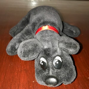May include: Black plush toy dog with large black eyes and a red ribbon around its neck. The dog is lying on its side with its head resting on the ground.