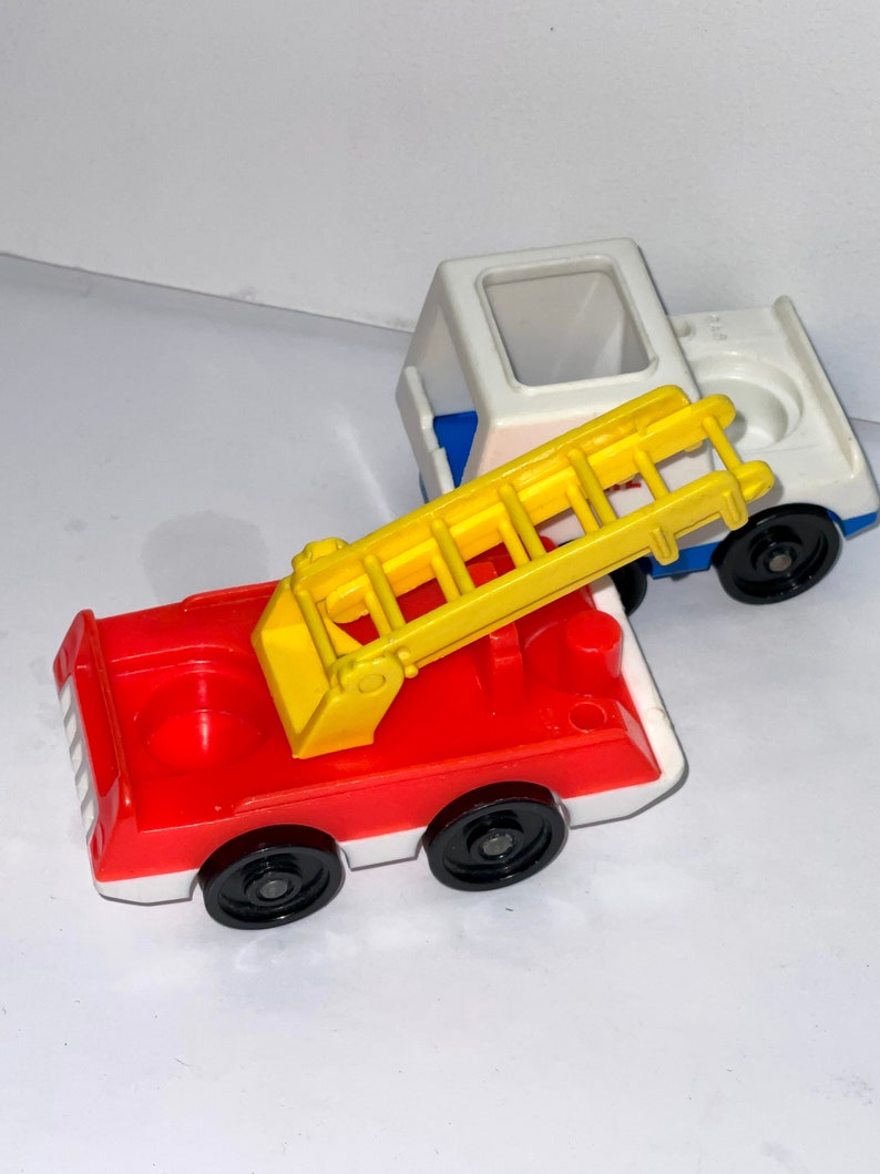 May include: A red fire truck with a yellow ladder and black wheels. The truck has a white cab and a red body. There is a white van with a blue cab and black wheels behind the fire truck.