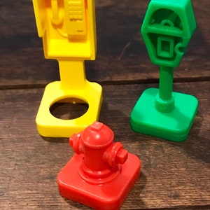 May include: Three plastic toy figurines: a yellow payphone, a green parking meter, and a red fire hydrant. The toys are on a wooden surface.