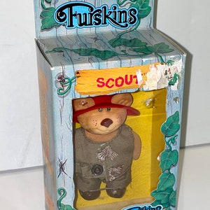 May include: A brown plush bear wearing a green vest and a red hat with a scout logo. The bear is packaged in a blue and yellow box with the words "Furskins" and "Scout" on it.