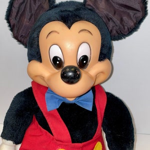 May include: A plush Mickey Mouse toy with black fur, brown ears, and a red and yellow jumpsuit. The toy has a blue bow tie and a friendly expression.