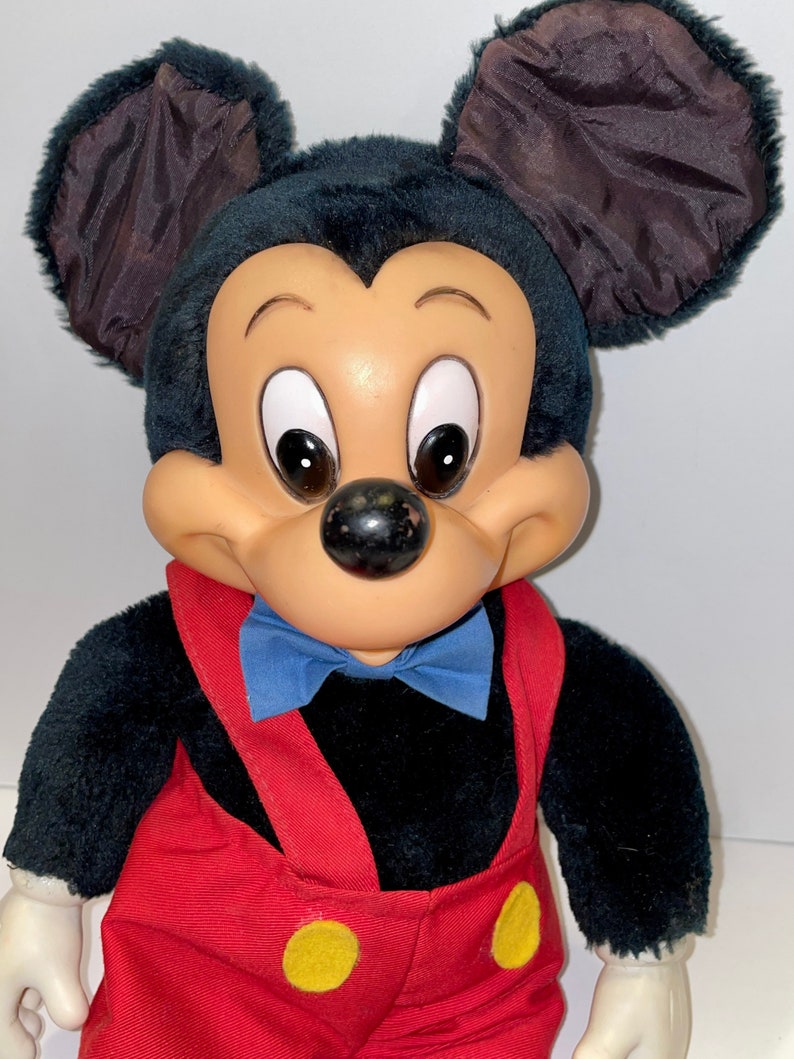 May include: A plush Mickey Mouse toy with black fur, brown ears, and a red and yellow jumpsuit. The toy has a blue bow tie and a friendly expression.