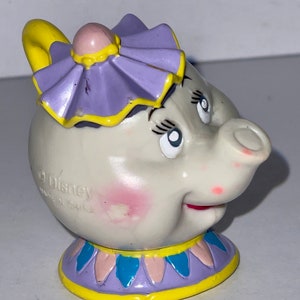 May include: A white ceramic teapot with a purple and yellow lid and a painted face. The teapot has a pink blush on its cheeks and a red mouth. The teapot is a character from the Disney movie Beauty and the Beast.