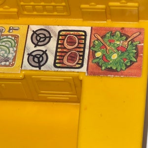 K&ouml;nnte beinhalten: Gelbes Spielzeug-K&uuml;chenset aus Plastik mit Herd, Sp&uuml;le und Schr&auml;nken. Der Herd hat zwei Kochfelder und einen Grill. Die Sp&uuml;le hat einen Wasserhahn und eine Sch&uuml;ssel mit gr&uuml;nem Gem&uuml;se. Die Schr&auml;nke haben T&uuml;ren, die sich &ouml;ffnen und schlie&szlig;en lassen.