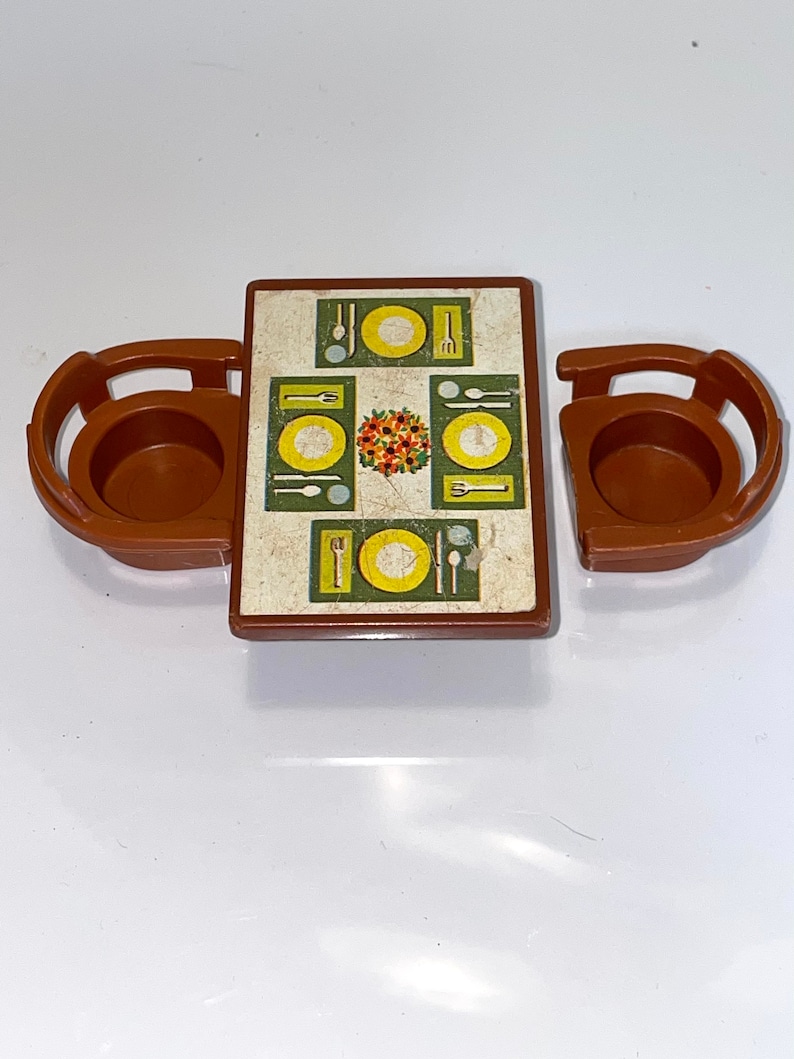 May include: A brown plastic toy table set with two chairs. The table has a green and yellow design with plates, forks, spoons, and a centerpiece.