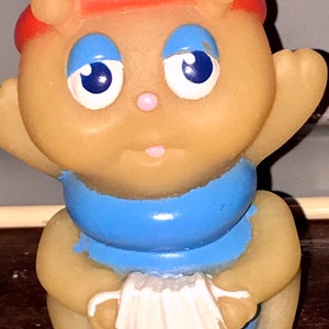 Puede incluir: Un peque&ntilde;o juguete de goma amarillo con un sombrero rojo, ojos azules y una bufanda azul. El juguete sostiene un acorde&oacute;n blanco.