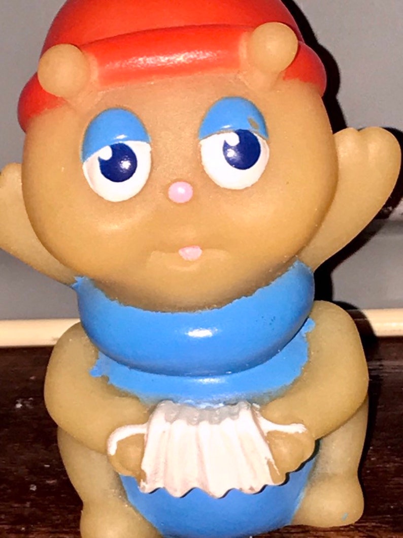 Puede incluir: Un peque&ntilde;o juguete de goma amarillo con un sombrero rojo, ojos azules y una bufanda azul. El juguete sostiene un acorde&oacute;n blanco.