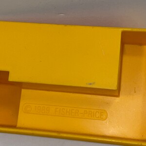 K&ouml;nnte beinhalten: Gelbes Spielzeug aus Kunststoff mit einem Copyright-Symbol und dem Text "1989 FISHER-PRICE" auf der Unterseite.