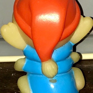 Puede incluir: Una peque&ntilde;a figura de pl&aacute;stico amarilla de una oruga que lleva un mono azul y un gorro de Pap&aacute; Noel rojo.