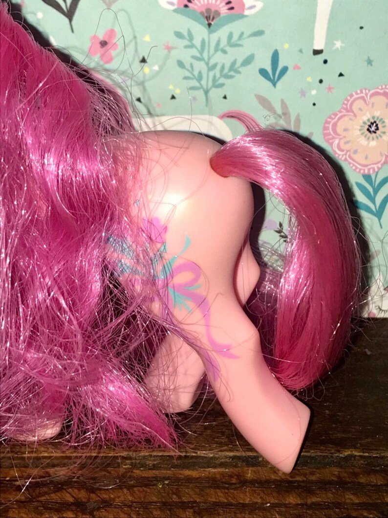 Puede incluir: Juguete de pony rosa con una larga melena y cola rosas. El pony tiene un dise&ntilde;o floral azul y morado en un lado.