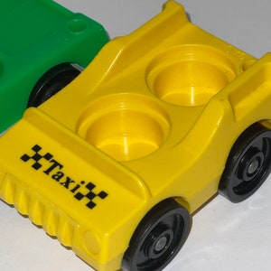May include: A yellow toy taxi with black wheels and a black "Taxi" sign on the front. The toy has two empty seats.