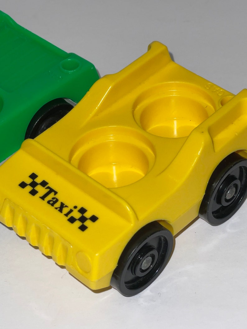May include: A yellow toy taxi with black wheels and a black "Taxi" sign on the front. The toy has two empty seats.