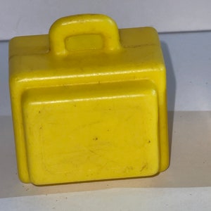 K&ouml;nnte beinhalten: Ein gelber Plastik-Spielzeugkoffer mit einem Griff oben. Der Koffer ist etwa 5 cm hoch und 7,5 cm breit.