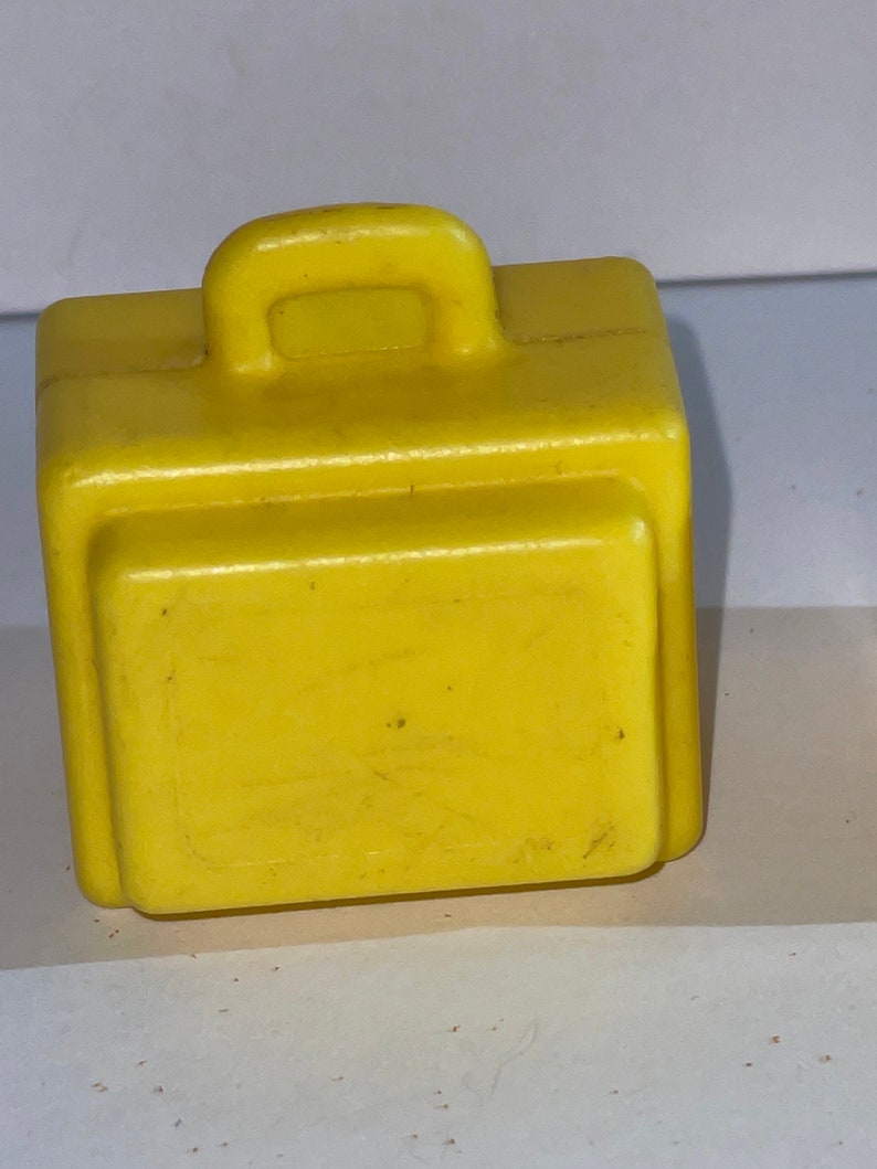 K&ouml;nnte beinhalten: Ein gelber Plastik-Spielzeugkoffer mit einem Griff oben. Der Koffer ist etwa 5 cm hoch und 7,5 cm breit.