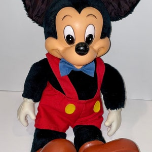 May include: A plush Mickey Mouse toy with black fur, red shorts, yellow buttons, and brown shoes. The toy has a blue bow tie and a friendly expression.