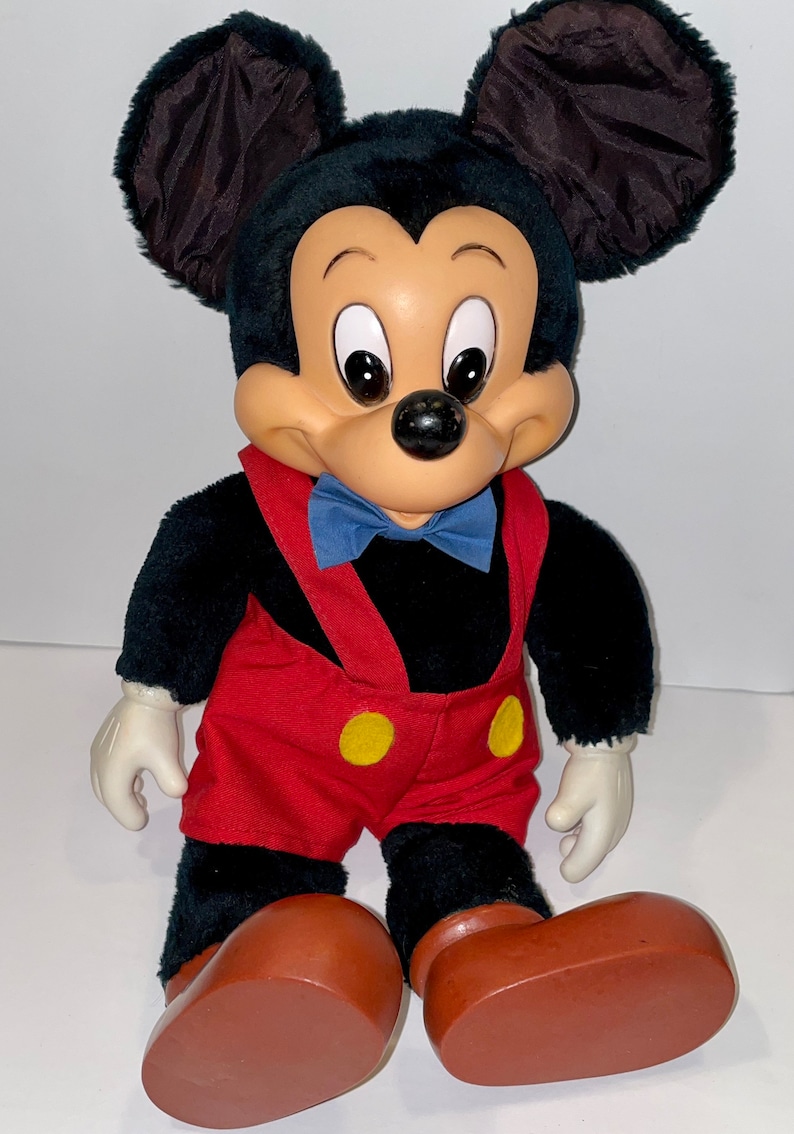 May include: A plush Mickey Mouse toy with black fur, red shorts, yellow buttons, and brown shoes. The toy has a blue bow tie and a friendly expression.