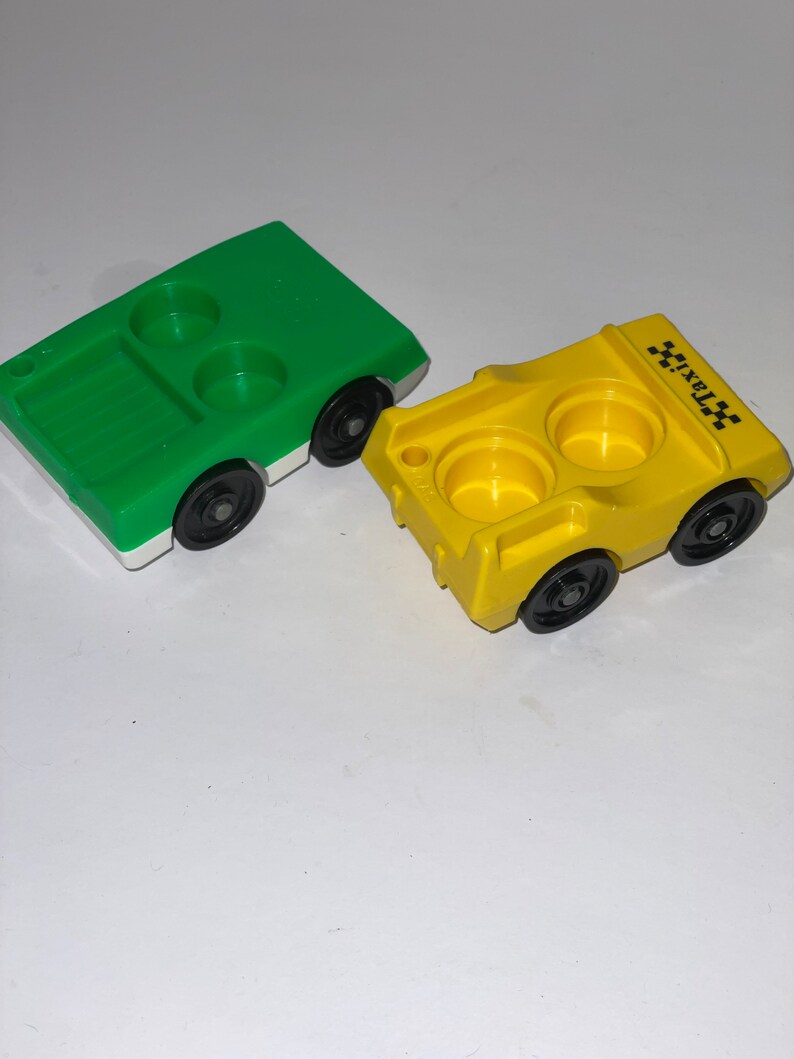 May include: Two plastic toy cars, one green and one yellow. The yellow car has "TAXI" printed on the side. Both cars have two cup holders.