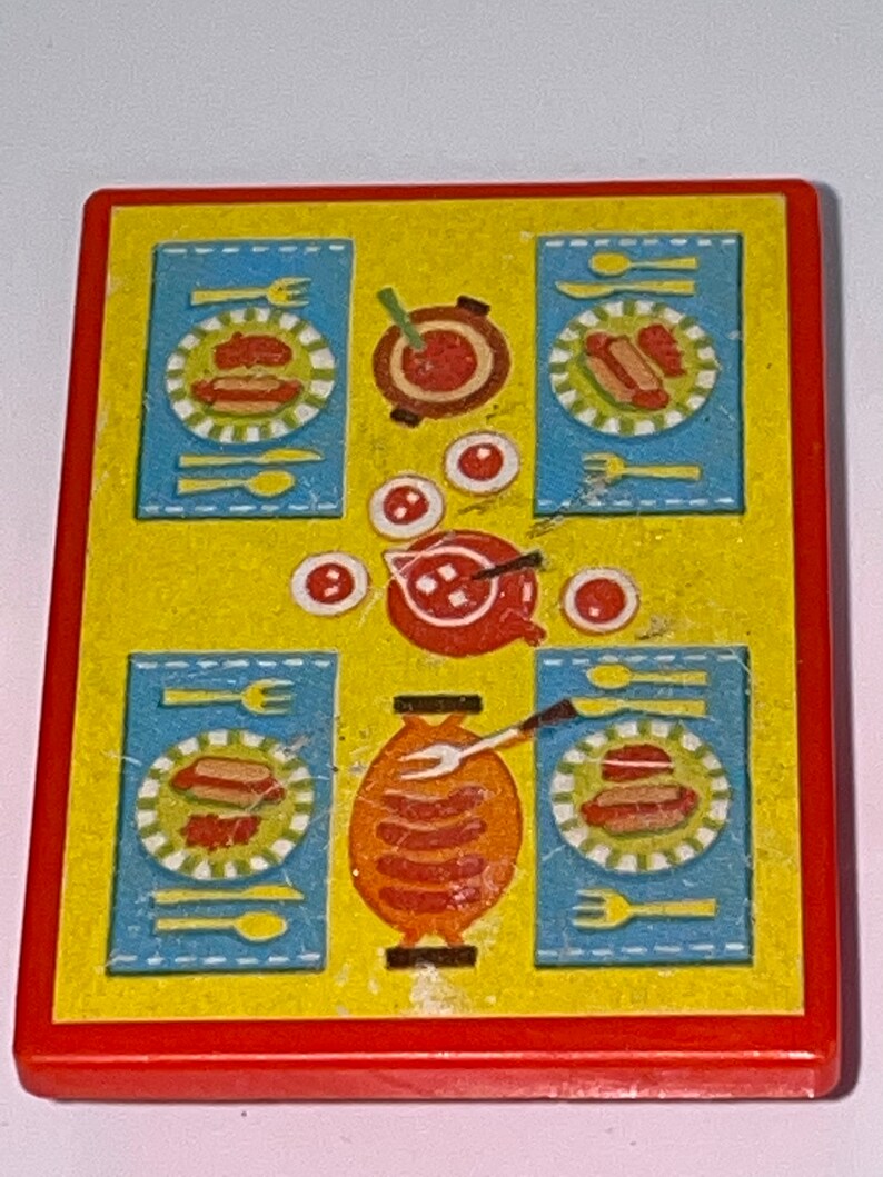 Pu&ograve; includere: Un tavolo da gioco in plastica giallo e rosso con una tovaglia a quadri blu e bianchi. Il tavolo ha una pentola rossa con manico, quattro piatti con hot dog e una griglia con salsicce.
