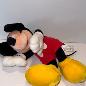 May include: A plush Mickey Mouse toy lying on its side with its head resting on its arm. The toy is wearing a red shirt with black outlines and yellow shoes. The toy has black eyes and a black nose.