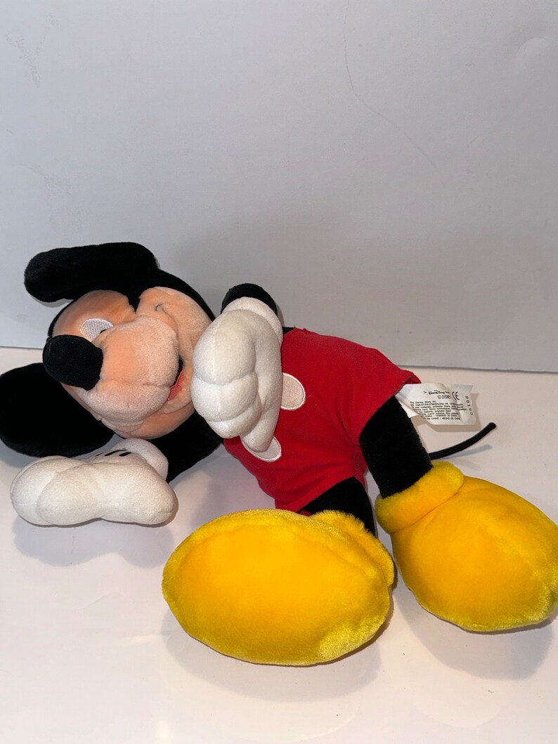 May include: A plush Mickey Mouse toy lying on its side with its head resting on its arm. The toy is wearing a red shirt with black outlines and yellow shoes. The toy has black eyes and a black nose.