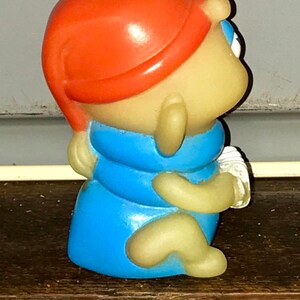 Puede incluir: Una peque&ntilde;a figura de pl&aacute;stico amarilla de un personaje que lleva un sombrero rojo y una camisa azul. El personaje est&aacute; sentado con las piernas cruzadas y los brazos cruzados.