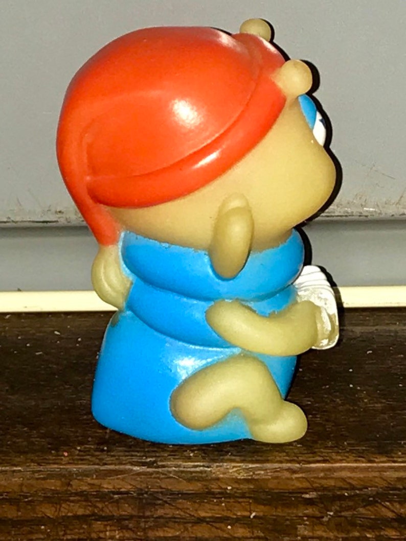 Puede incluir: Una peque&ntilde;a figura de pl&aacute;stico amarilla de un personaje que lleva un sombrero rojo y una camisa azul. El personaje est&aacute; sentado con las piernas cruzadas y los brazos cruzados.