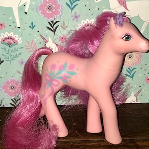 Puede incluir: Pony rosa My Little Pony con una flor morada en el pelo y un dise&ntilde;o floral en el costado. El pony tiene una melena y una cola rosas.