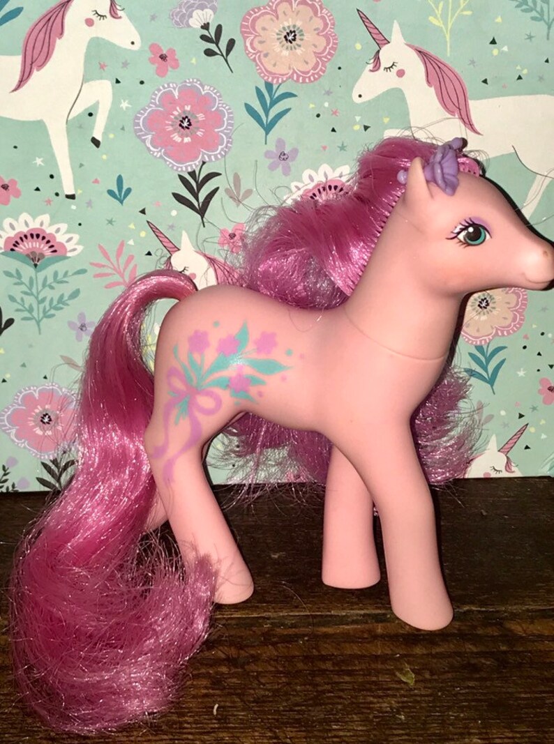 Puede incluir: Pony rosa My Little Pony con una flor morada en el pelo y un dise&ntilde;o floral en el costado. El pony tiene una melena y una cola rosas.