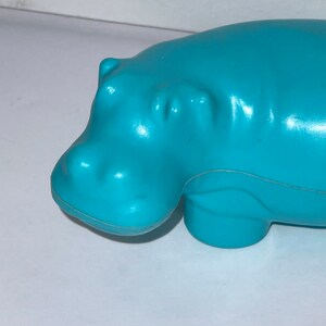 May include: A turquoise blue plastic hippopotamus toy. The toy is in a lying down position with its head turned to the left.