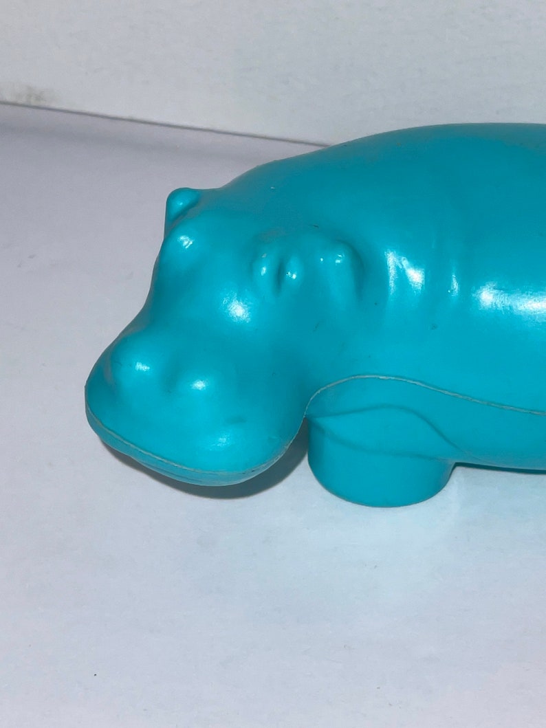 May include: A turquoise blue plastic hippopotamus toy. The toy is in a lying down position with its head turned to the left.