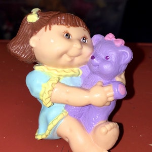 May include: A small plastic figurine of a Cabbage Patch Kid doll holding a purple teddy bear. The doll has brown hair, blue eyes, and is wearing a blue and yellow dress. The teddy bear has a pink bow on its head.